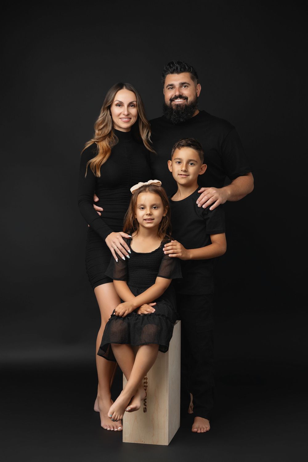 Studio family portrait with everyone wearing coordinated black outfits against dark background.