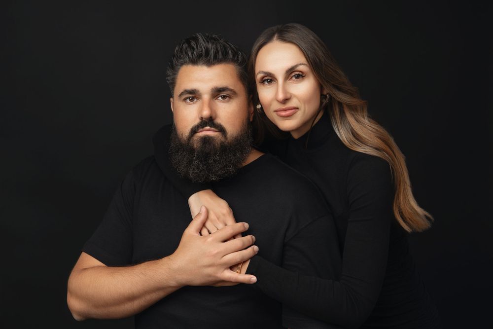 Couple in black turtlenecks pose for intimate portrait against dark background.
