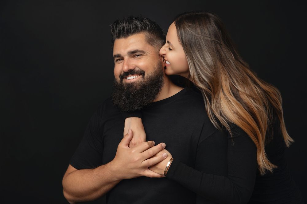 Romantic couple embraces against dark background, wearing black, sharing an intimate moment.