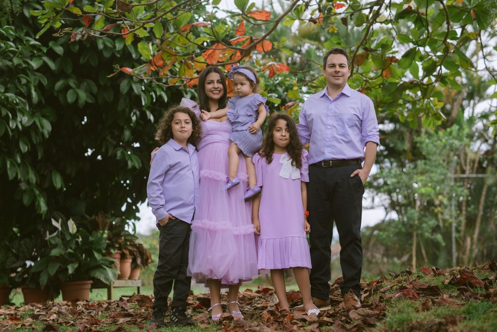 A family wearing coordinated purple and lavender outfits poses together in front of leafy trees during an outdoor portrait session.