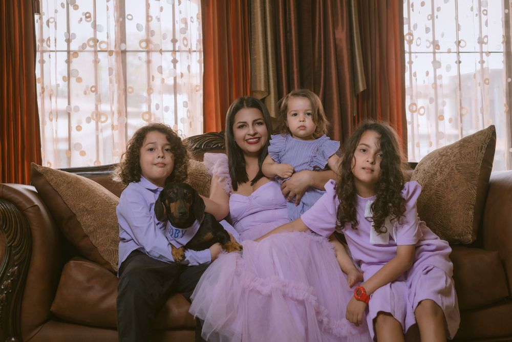 Four people in matching lavender outfits sit together on a brown sofa near lace curtained windows.