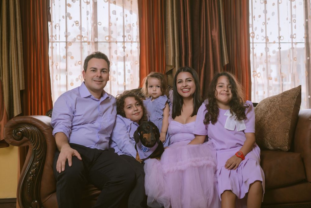 A family in coordinating purple and lavender outfits sits together with their dog on a brown couch by lace curtained windows.