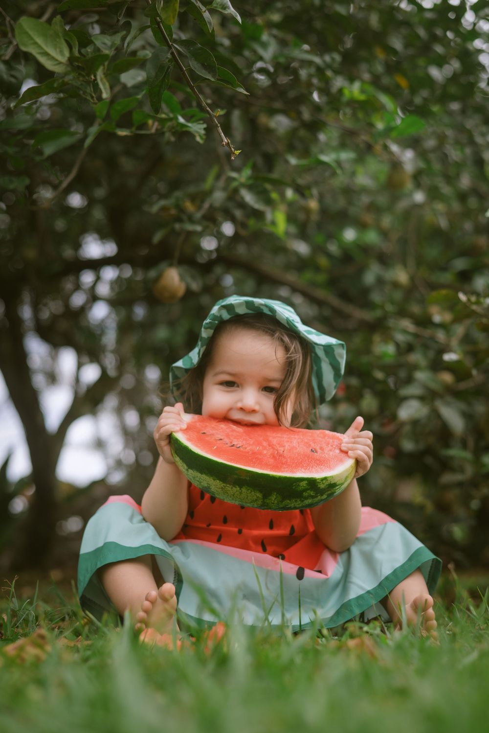 Little one in a green dress and bonnet enjoys a fresh slice of watermelon while sitting on grass under trees.