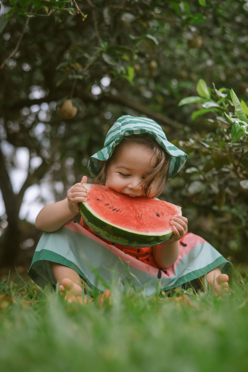 A small child in a green outfit and sunhat enjoys a large slice of watermelon while sitting in the grass.