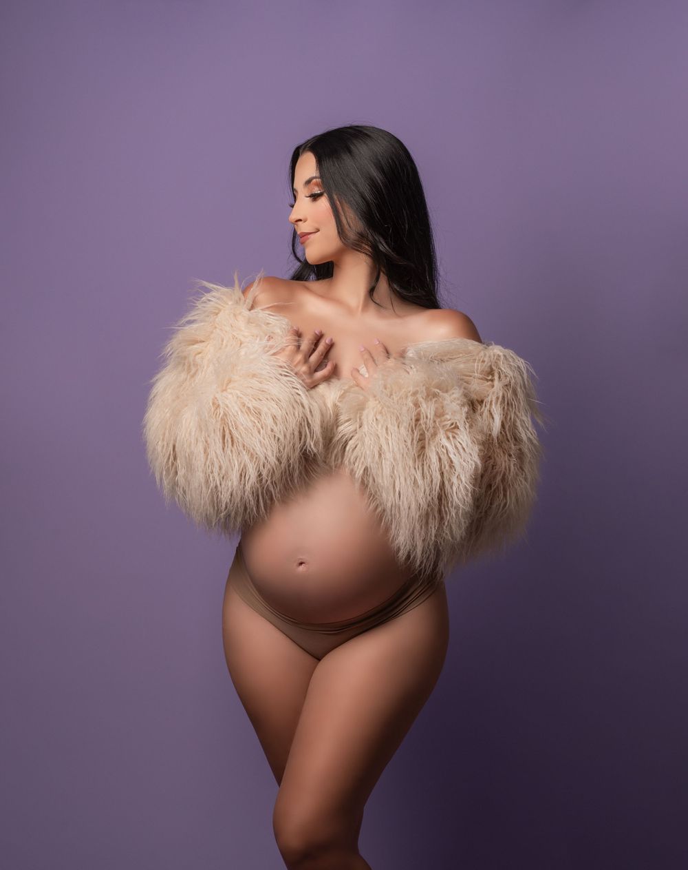 Artistic maternity photo with flowy beige fur wrap against purple backdrop in studio setting.