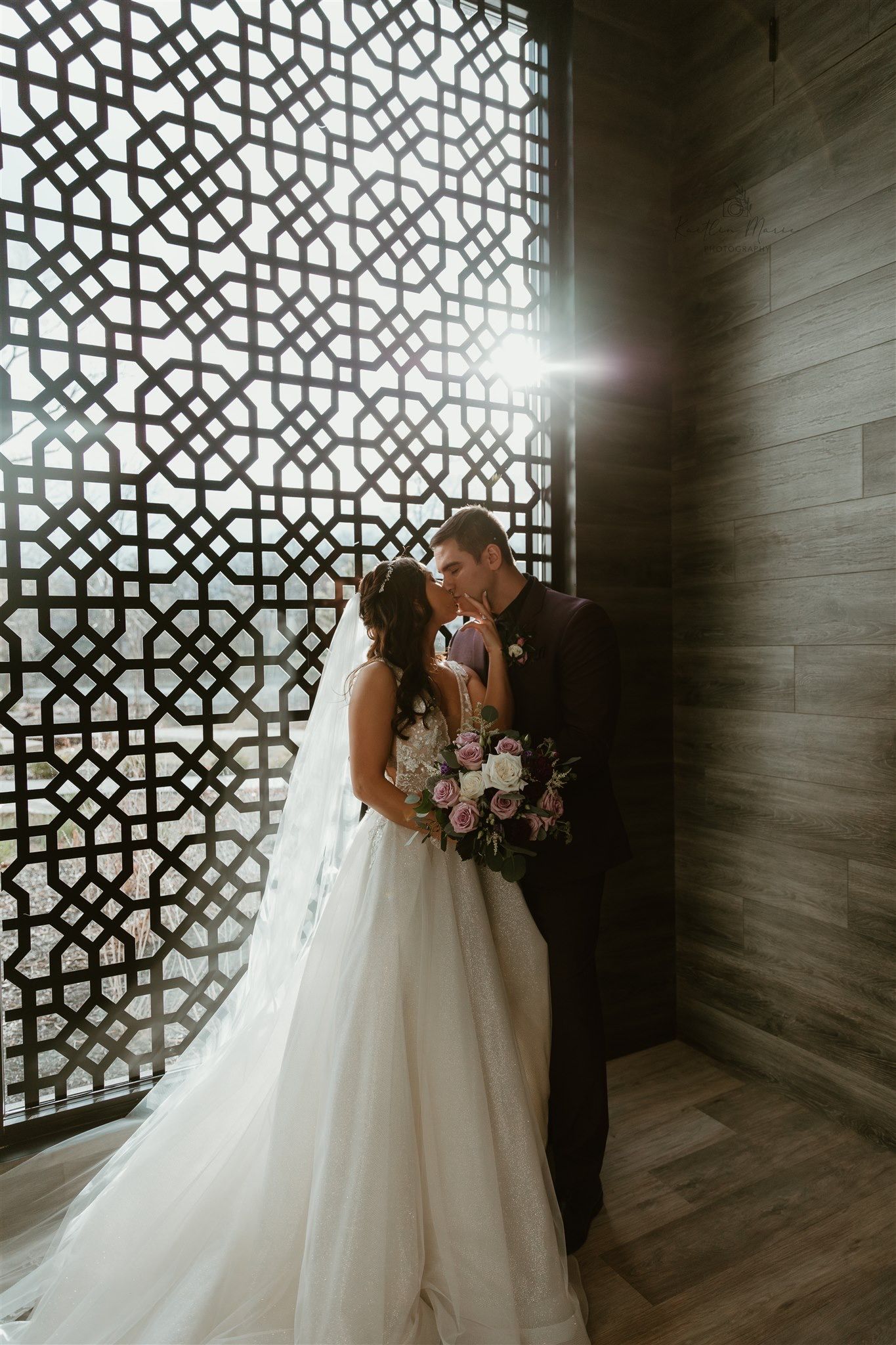 Wedding couple shares intimate moment in front of geometric patterned window with dramatic lighting.