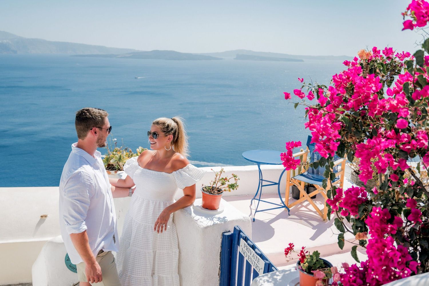 Couple enjoying drinks on white terrace with pink bougainvillea flowers overlooking Mediterranean Sea in Santorini.