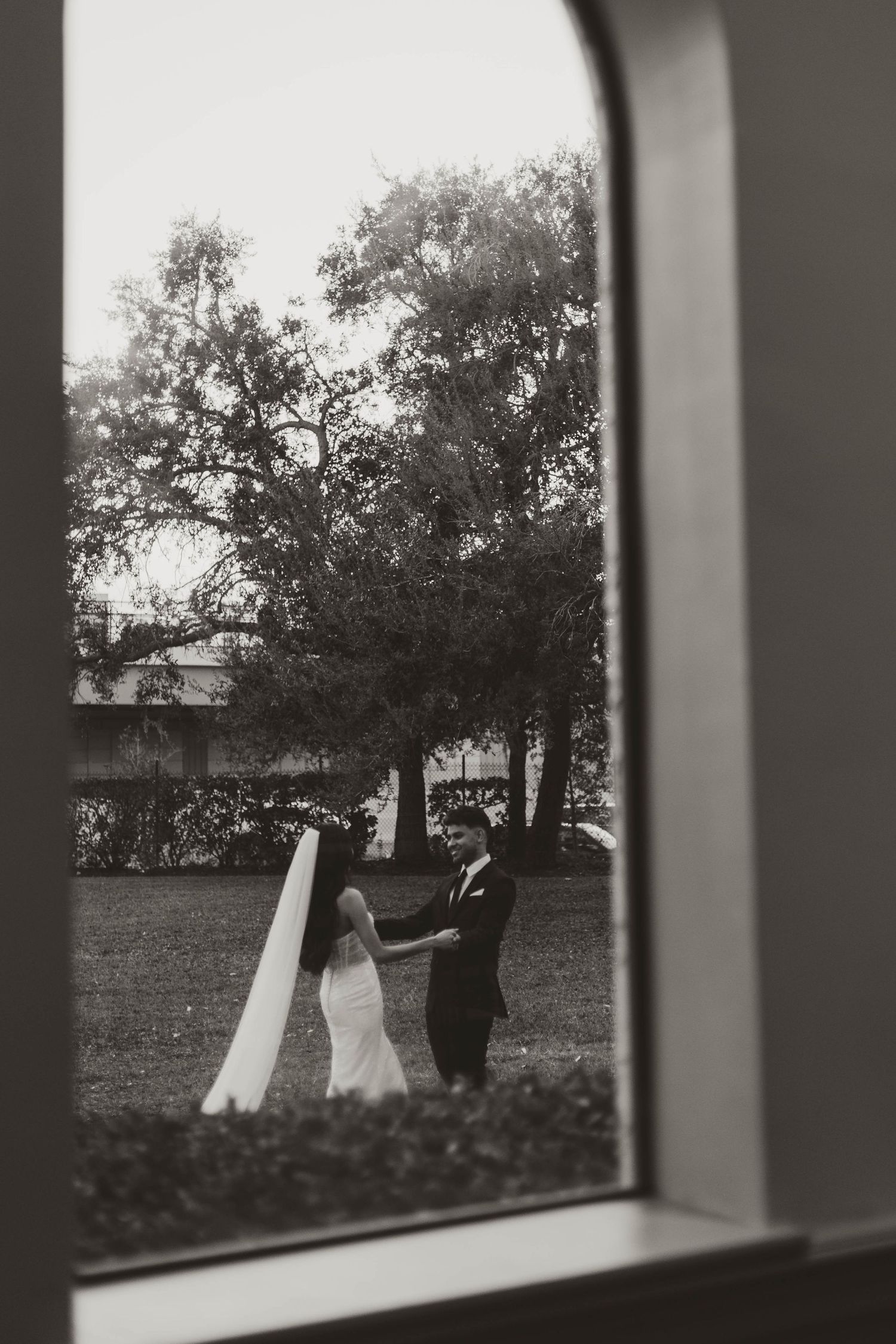 Black and white wedding photo framed through an elegant window showing ceremony on the lawn.