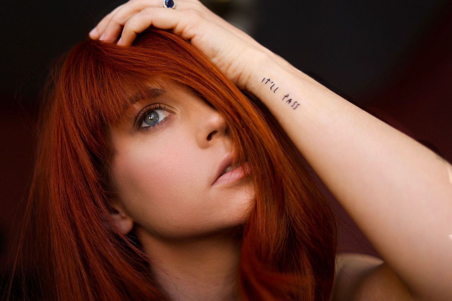 Dramatic portrait showcasing vibrant red hair and visible arm tattoo against dark background.