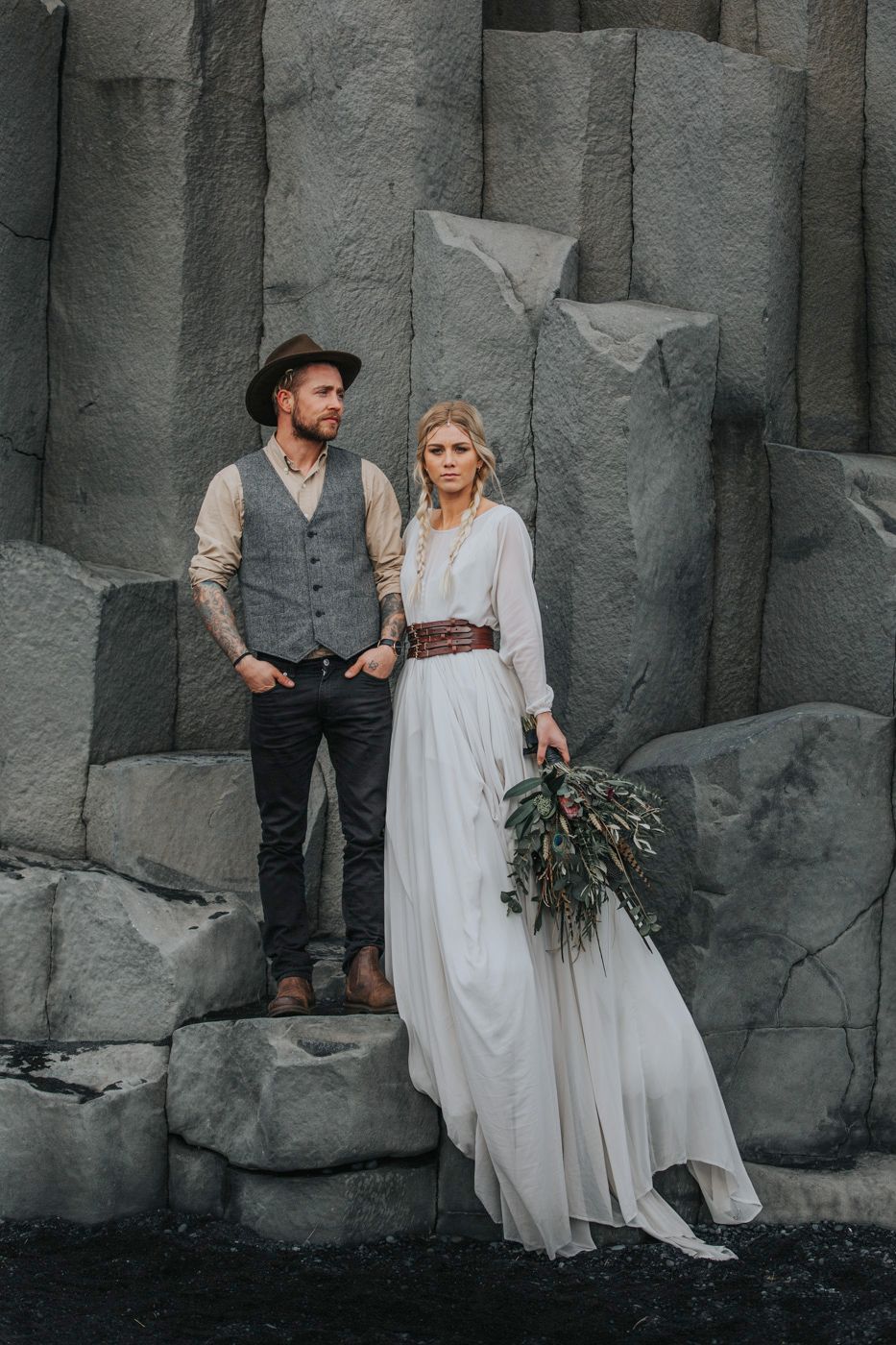 Couple in rustic wedding attire standing on dramatic basalt rock formations.