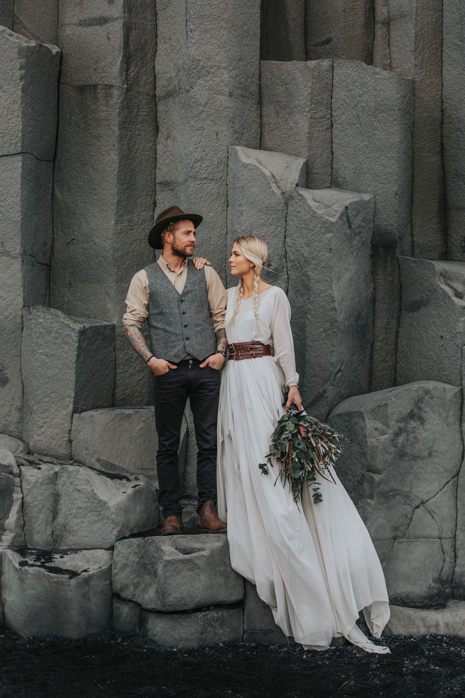 A bohemian wedding couple poses against geometric basalt cliffs with a natural bouquet.