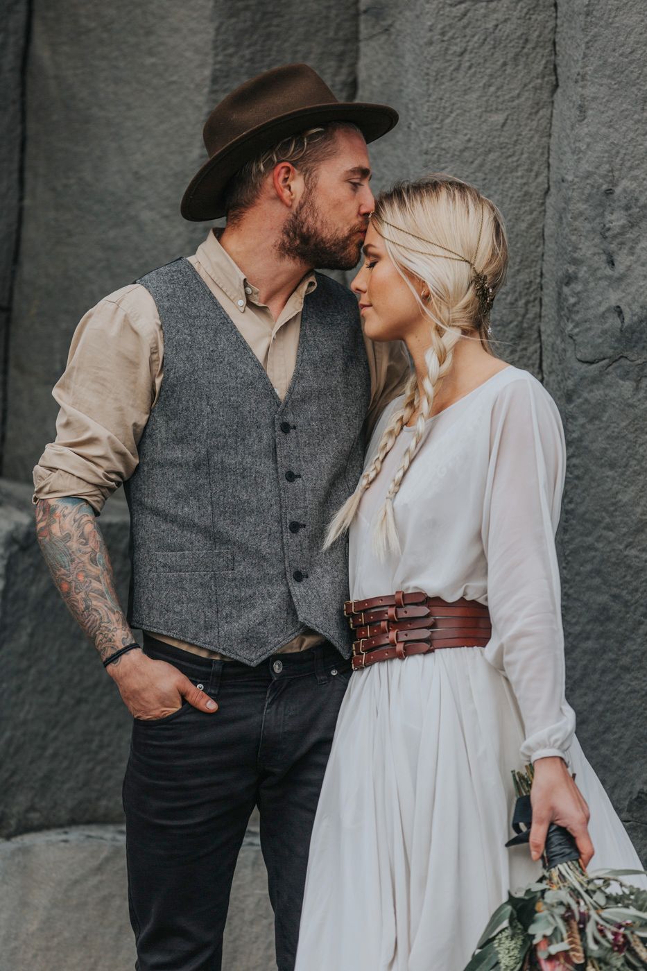 A romantic couple in vintage-inspired wedding attire pose against a grey stone wall with a rustic aesthetic.