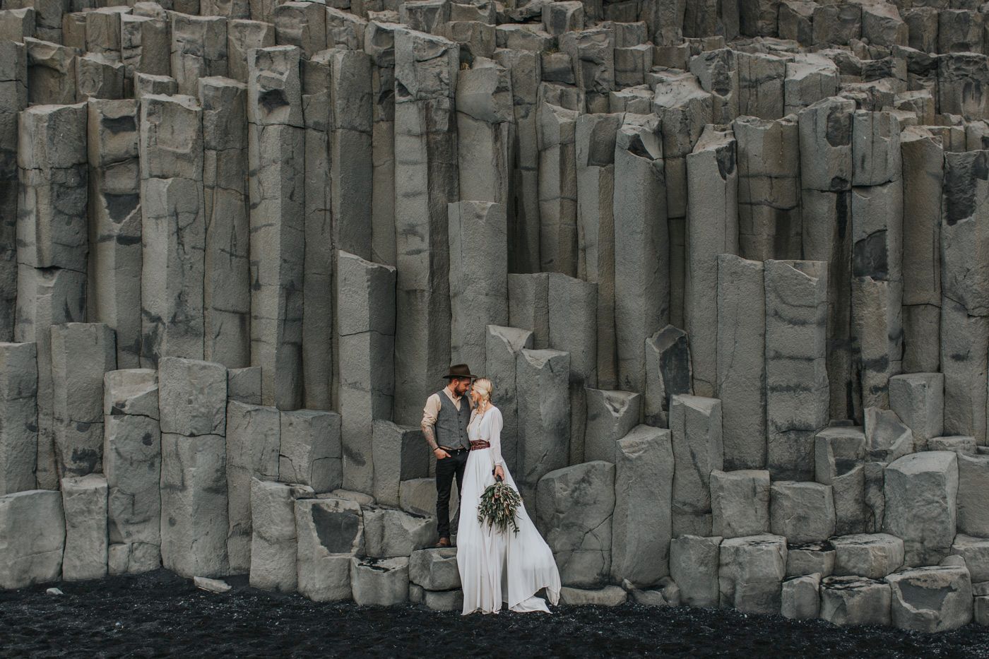 A wedding couple stands against dramatic basalt columns on a black sand beach.