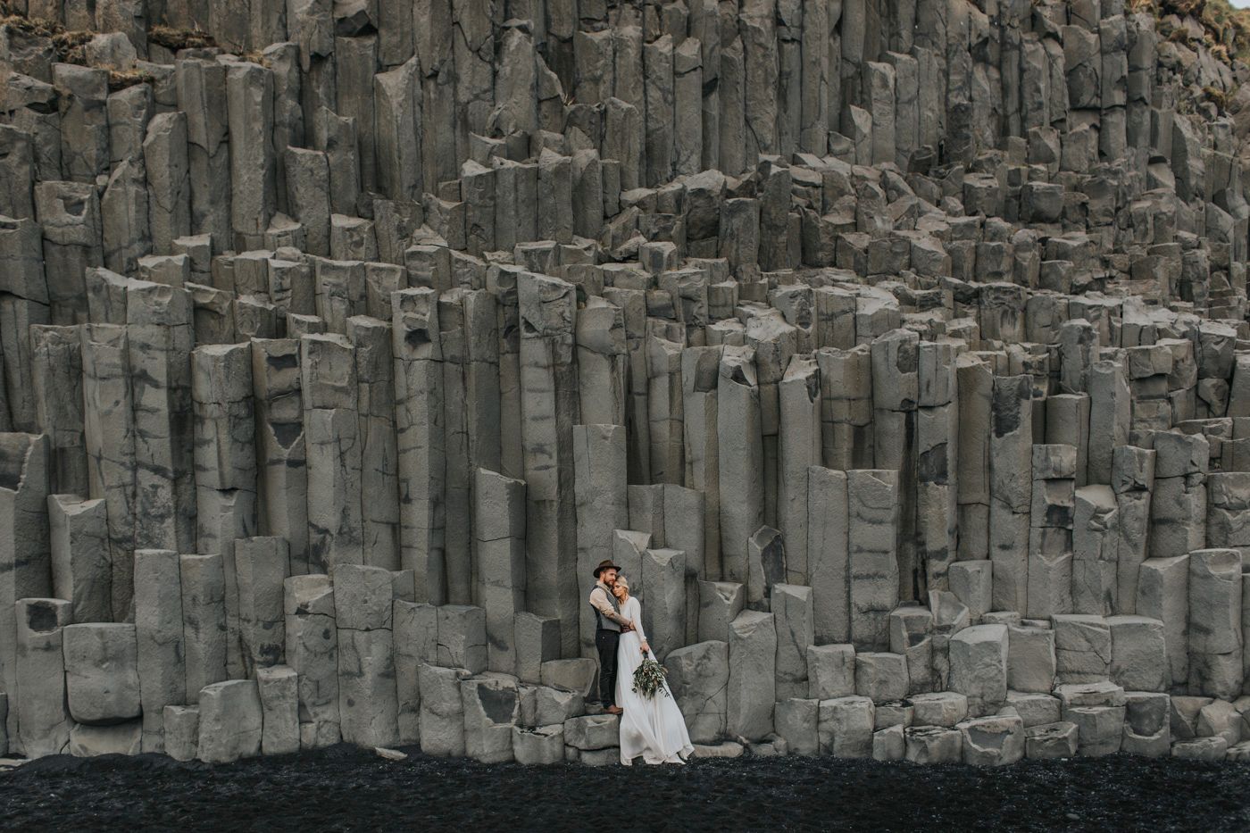 A tiny couple embraces against massive geometric basalt columns in Iceland.