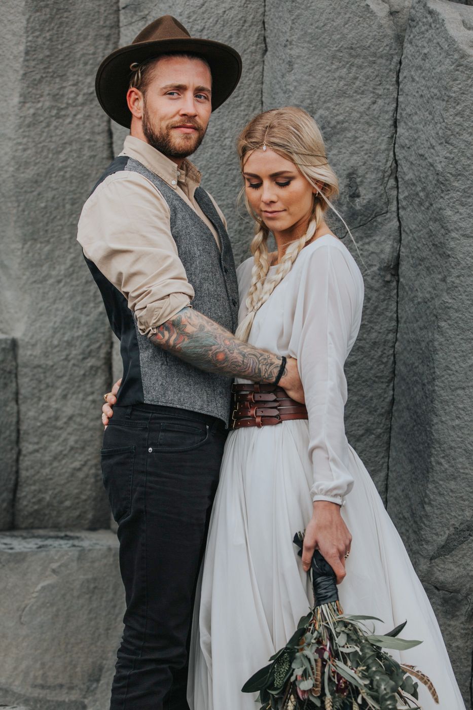A stylish couple in rustic wedding attire poses against a stone wall backdrop.