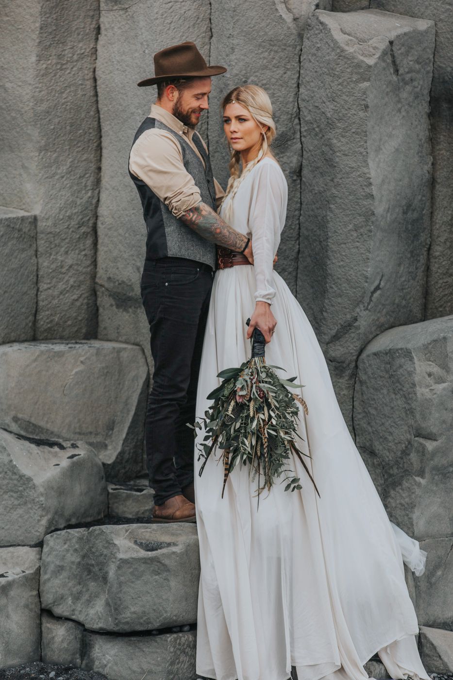Romantic wedding couple poses on dramatic rock formations with bride holding rustic greenery bouquet against grey stone wall.