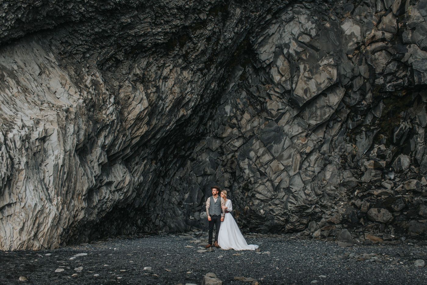 Wedding couple stands against dramatic black rocky cliff backdrop on dark beach.