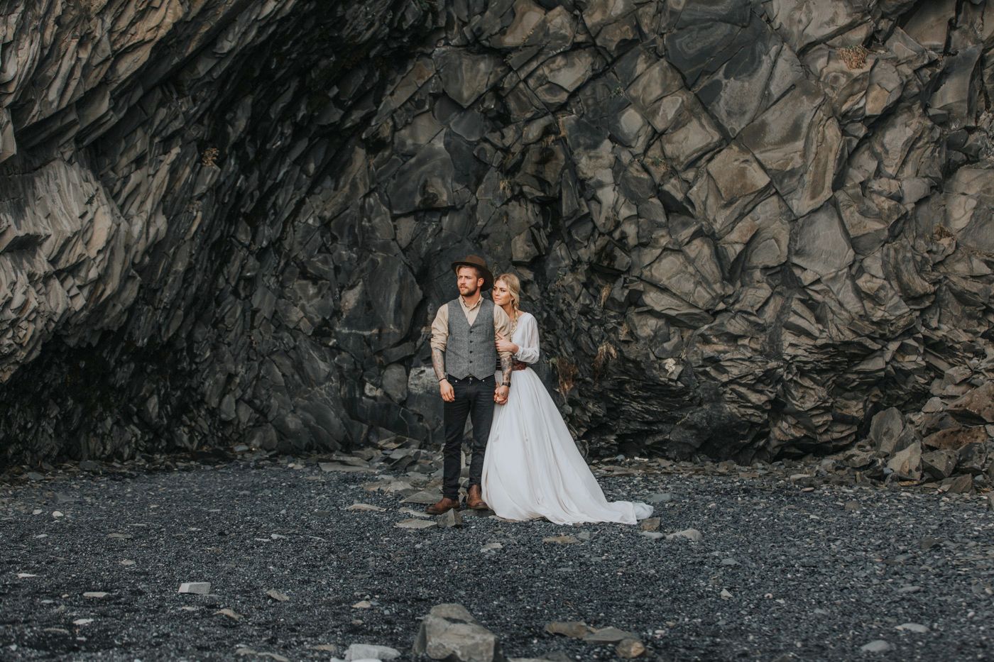 Wedding portrait with couple standing on black pebble beach in front of towering rock face.