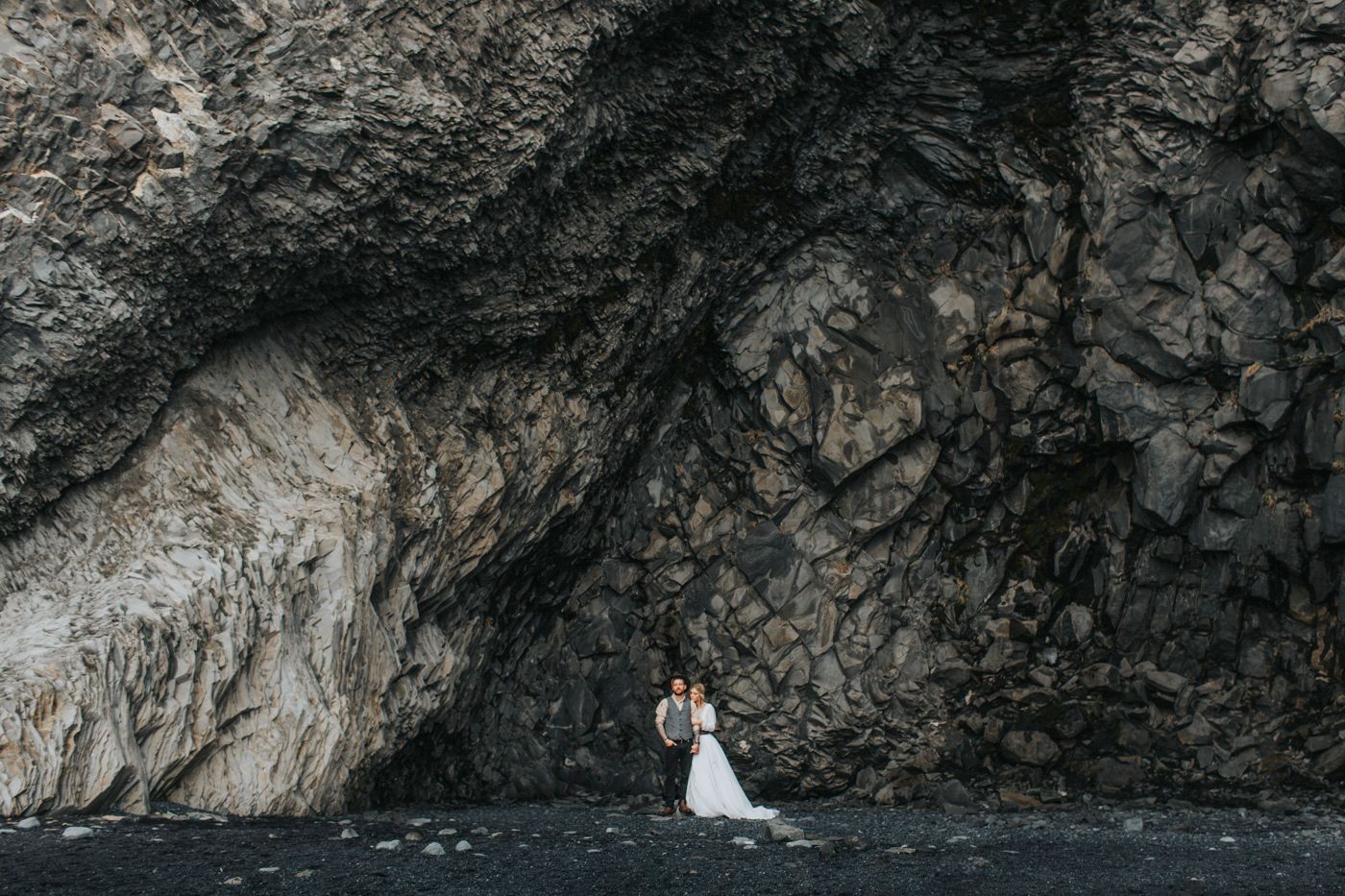 Wide shot of wedding couple appearing tiny against massive rocky cliff face.