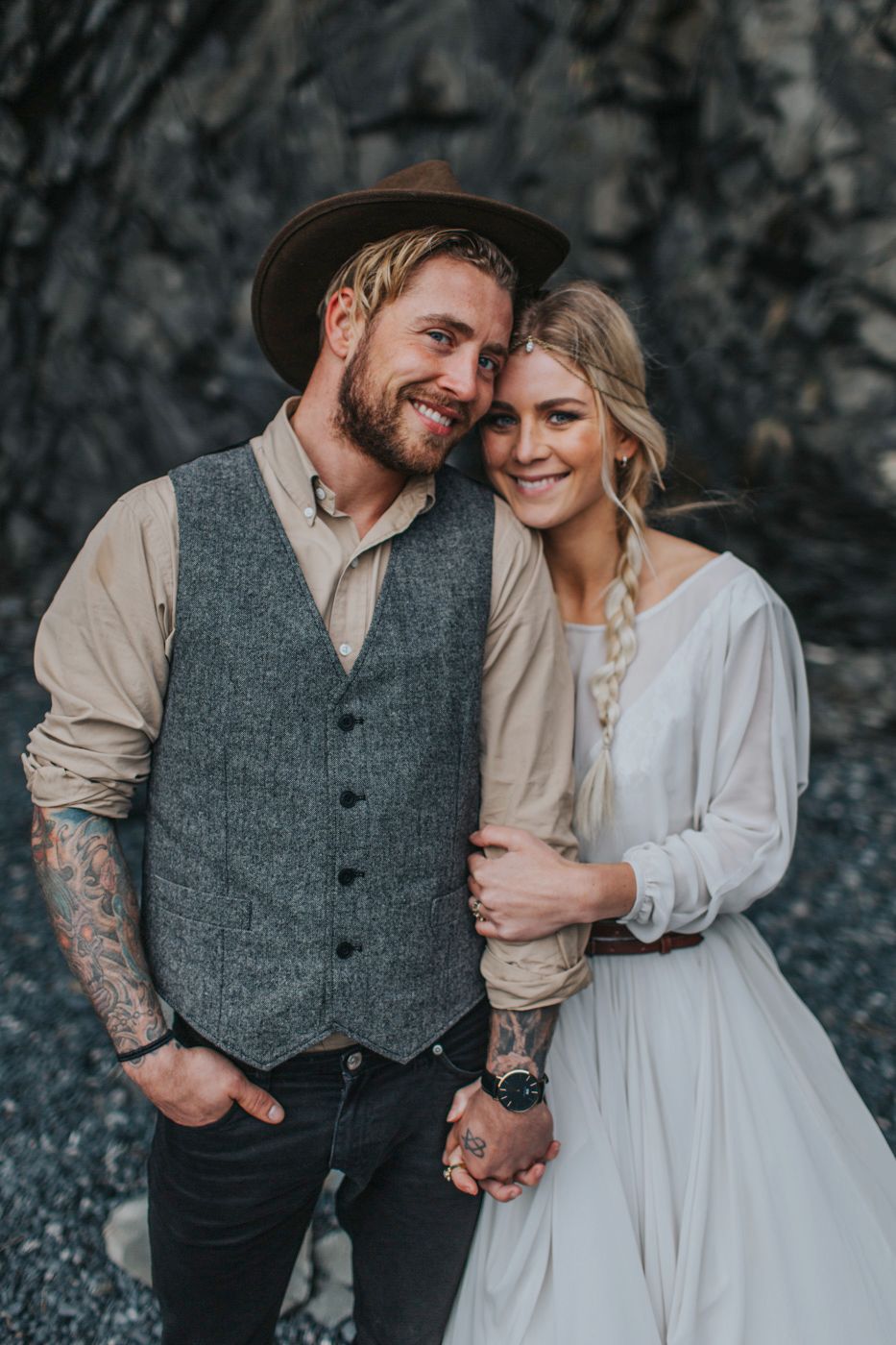 Rustic wedding couple poses against rocky wall with groom in vest and hat and bride in flowing white dress.
