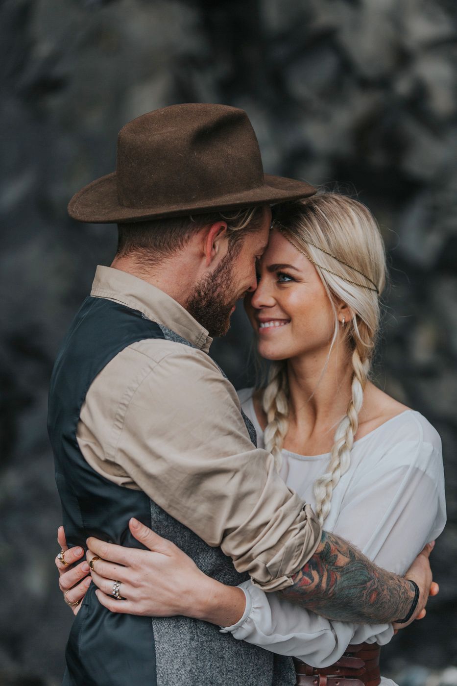 Tender embrace of newlyweds in rustic attire against dramatic stone wall.