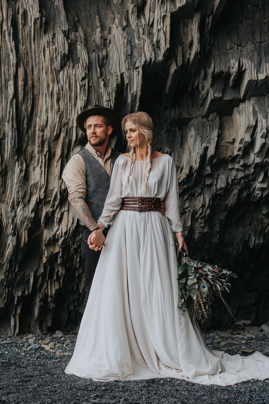 Wedding couple stands before dramatic rocky cliff face in rustic elegant attire.