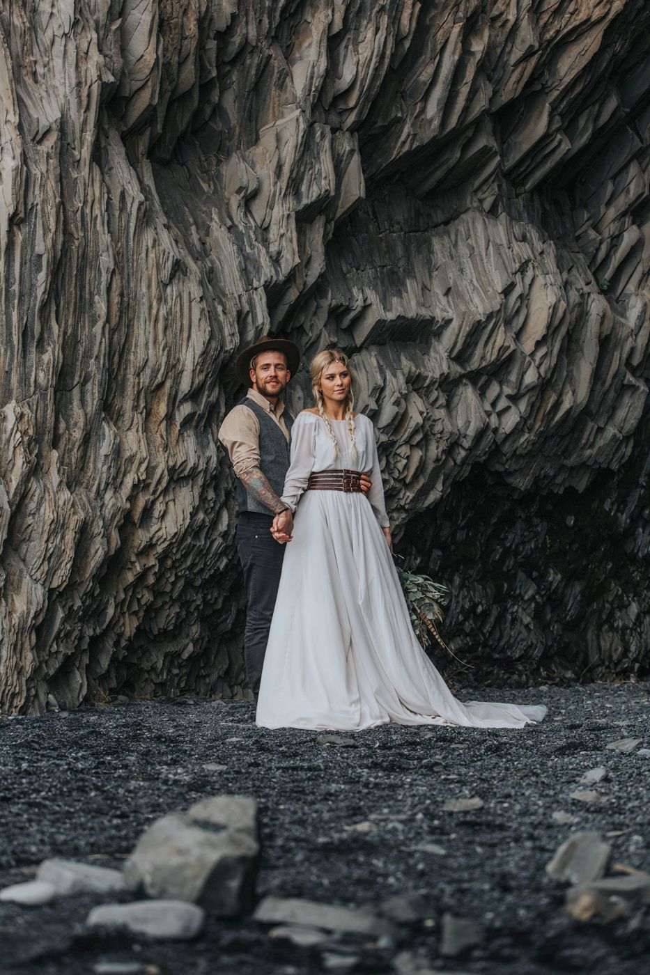 Wedding portrait featuring couple in rustic attire against dramatic cave-like rock formation.