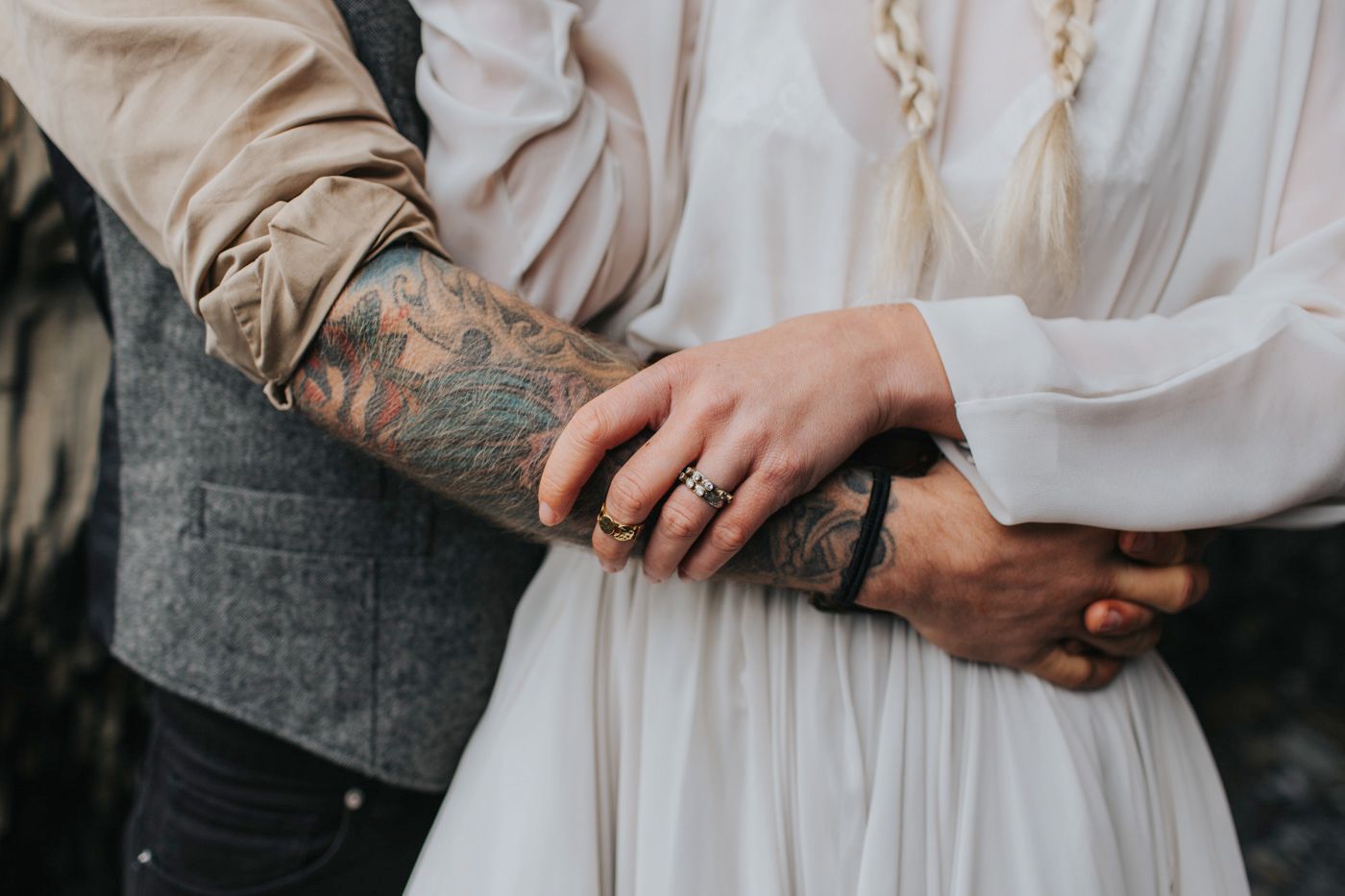Close-up shot of tattooed arms embracing in formal wedding attire against a light background.