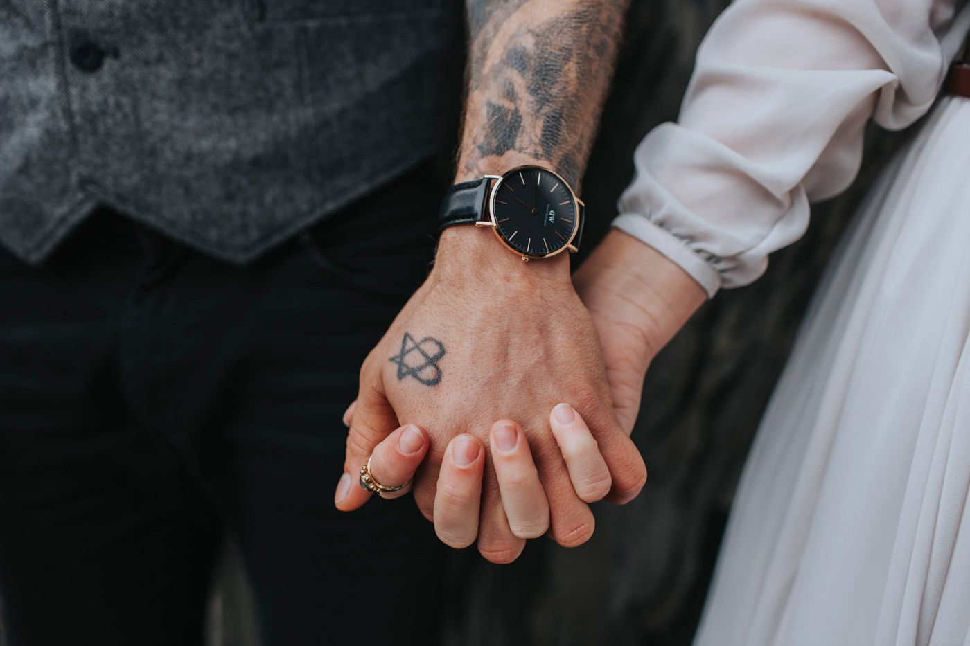 Close-up of hands with tattoo and watch holding each other against white fabric.