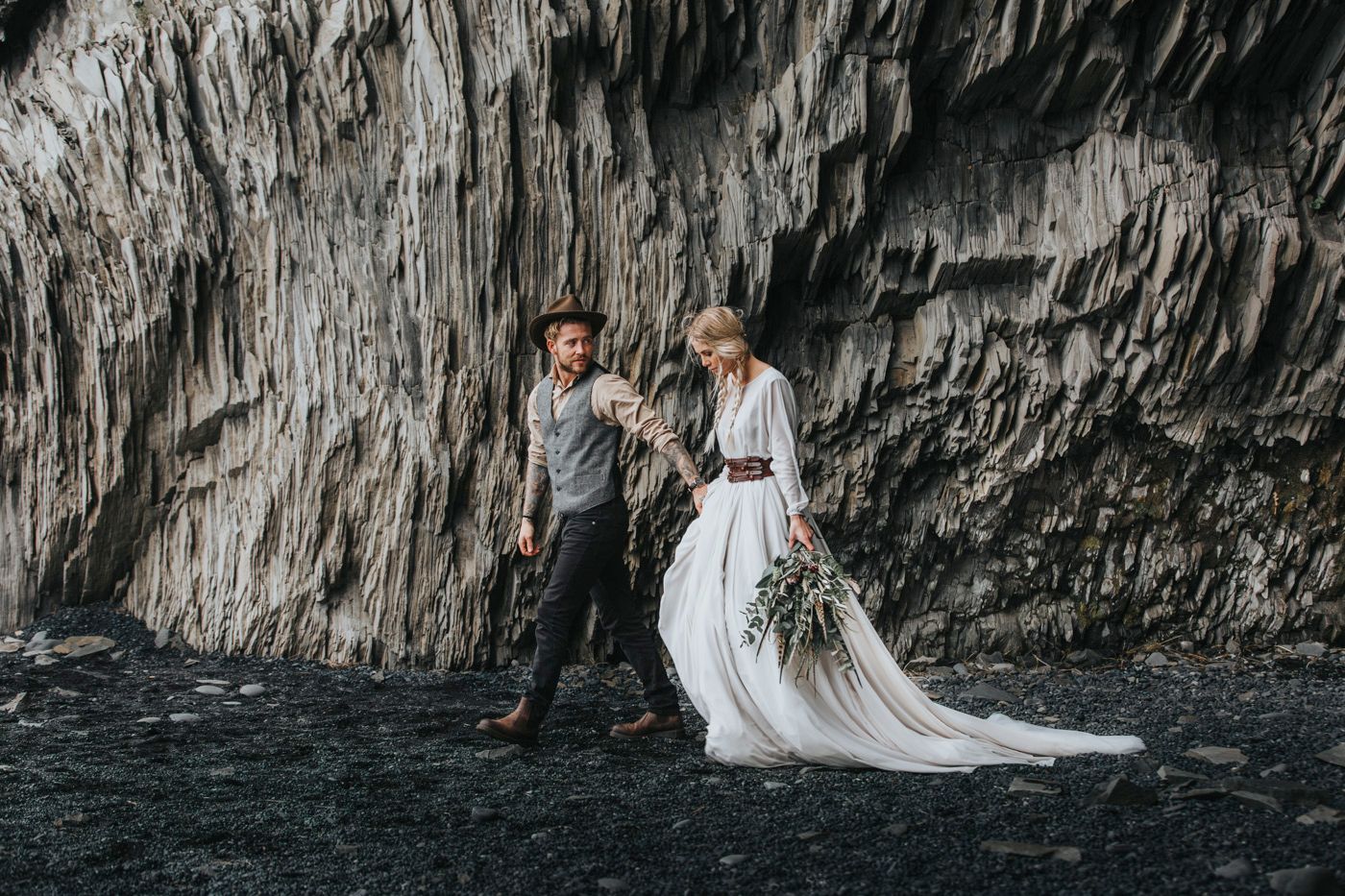 Elegant wedding couple posing on black volcanic beach near towering cliff face.