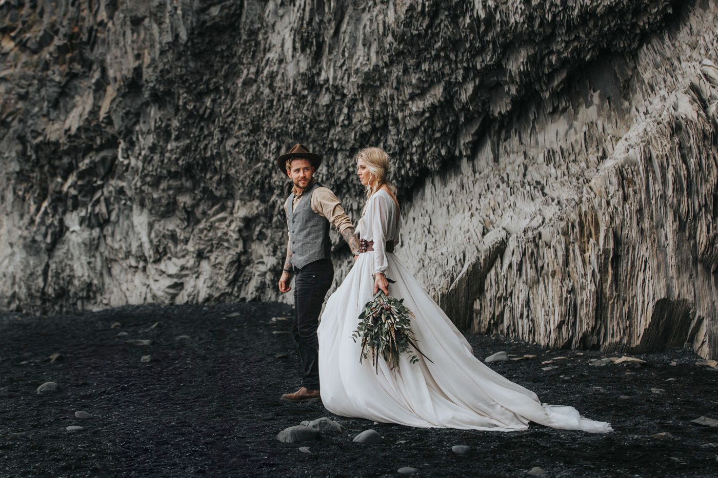 Wedding couple standing on black sand beach near dramatic volcanic cliffs.