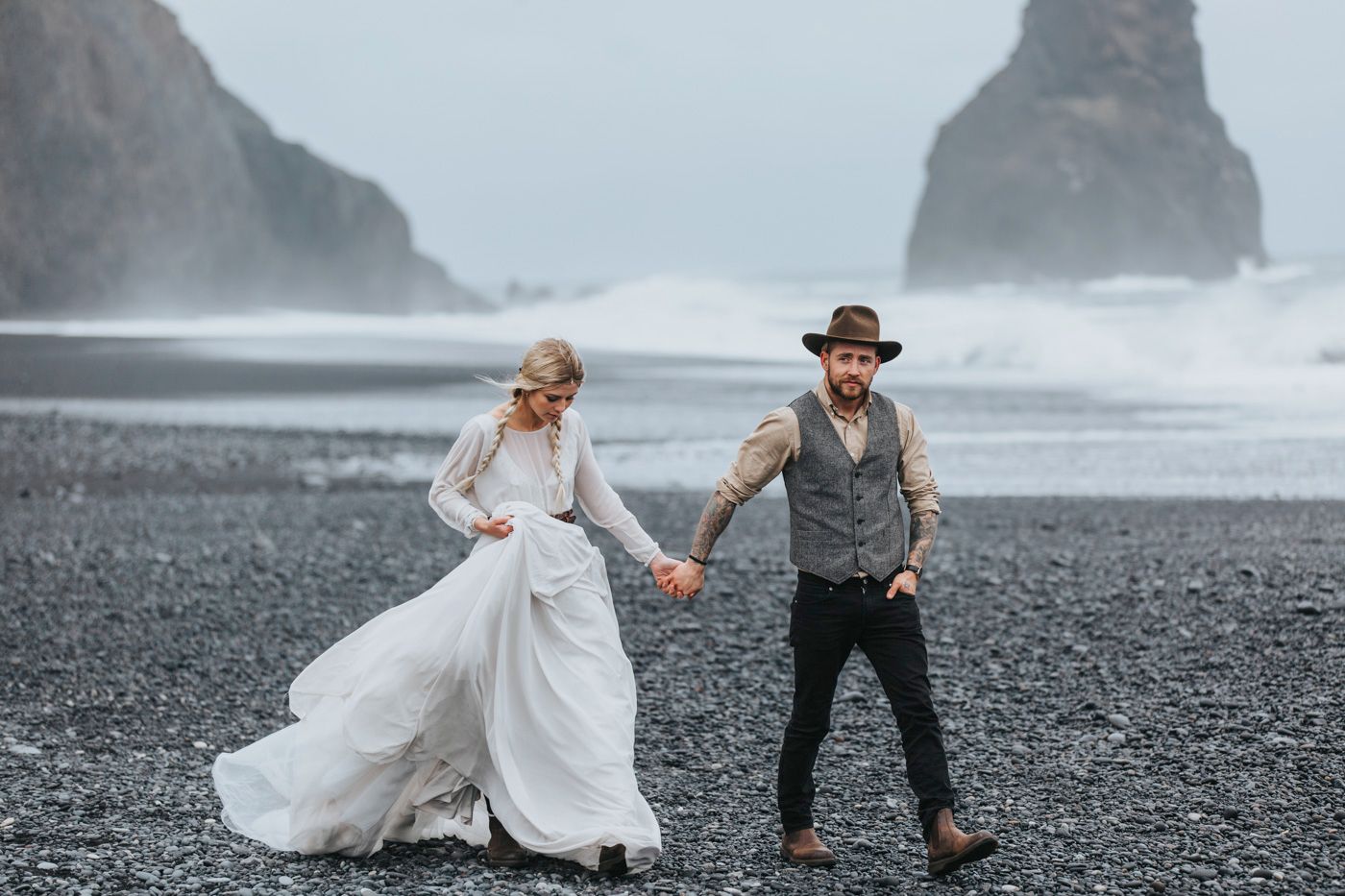 Couple in wedding attire walking hand in hand on dramatic black sand beach.