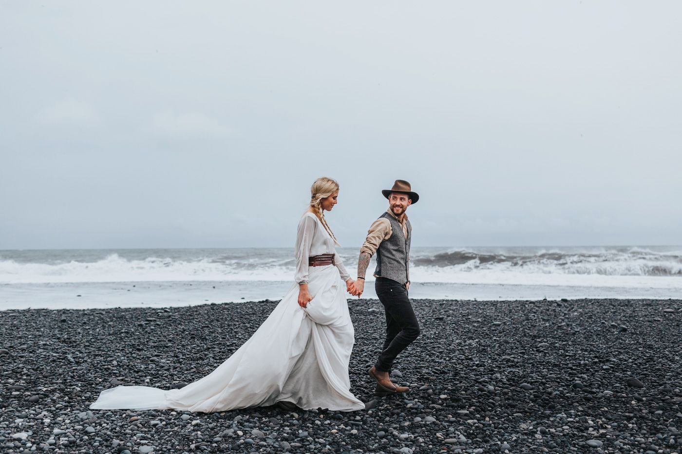 Couple in wedding attire dancing on dramatic black sand beach.
