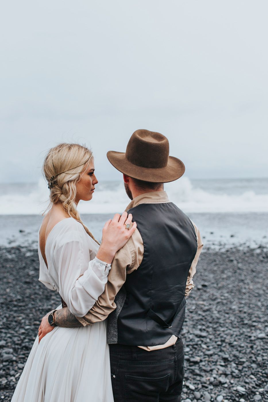 Couple in vintage-inspired wedding attire share an intimate moment on a dramatic black sand coastline.