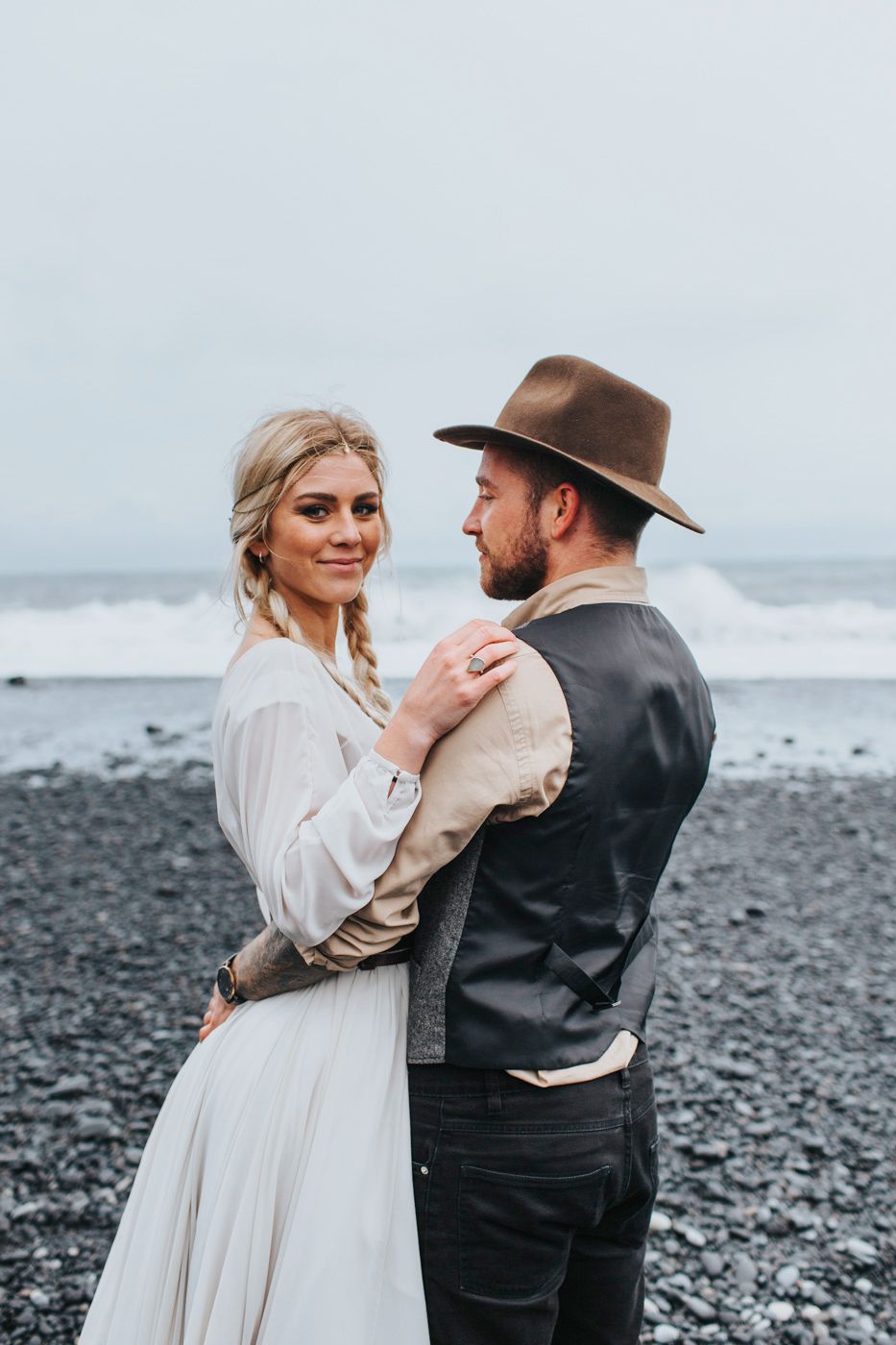 Wedding couple in rustic attire pose together on volcanic beach with waves crashing behind them.