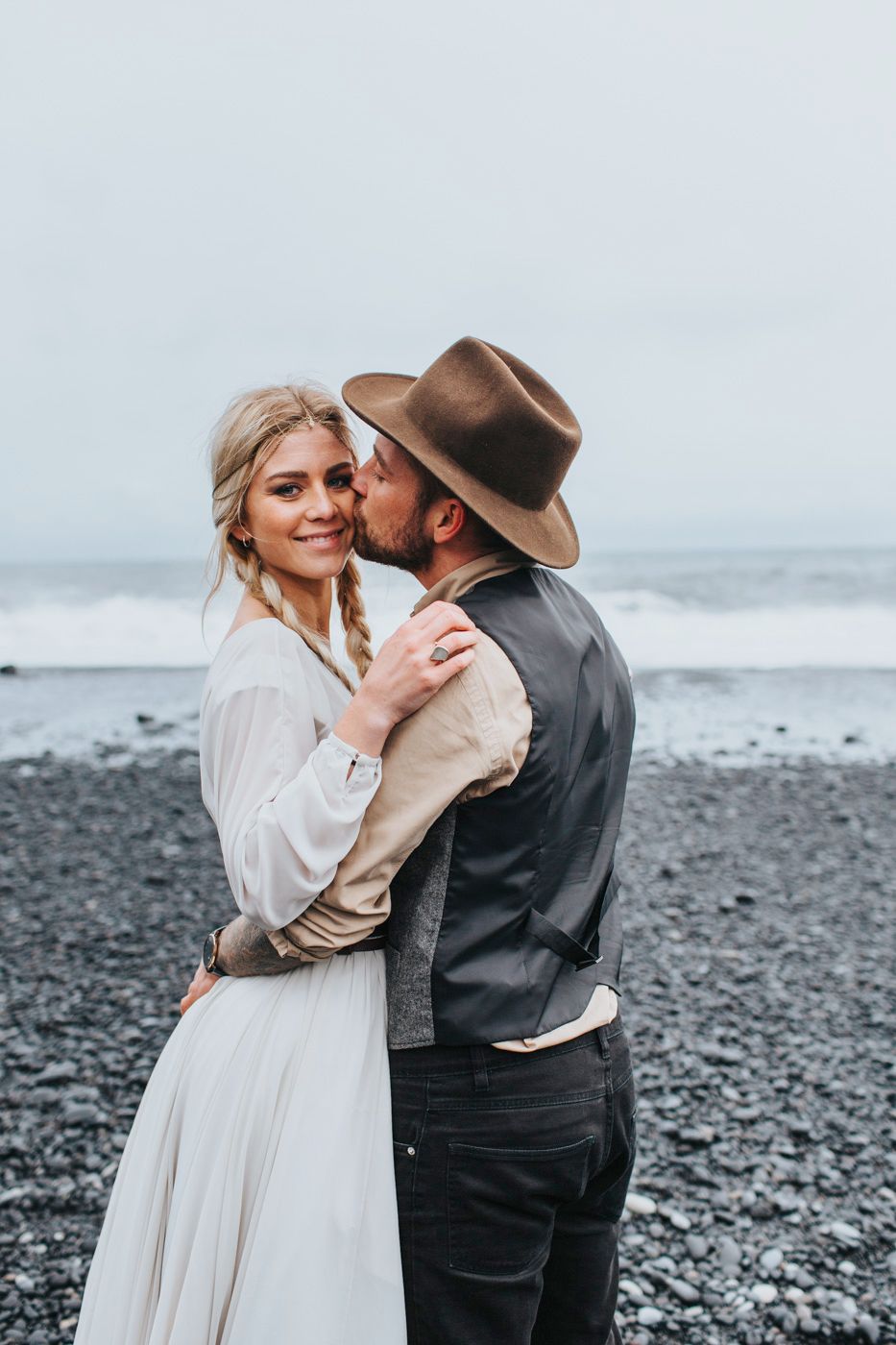 Joyful wedding couple shares tender embrace on dramatic black sand beach with ocean backdrop.