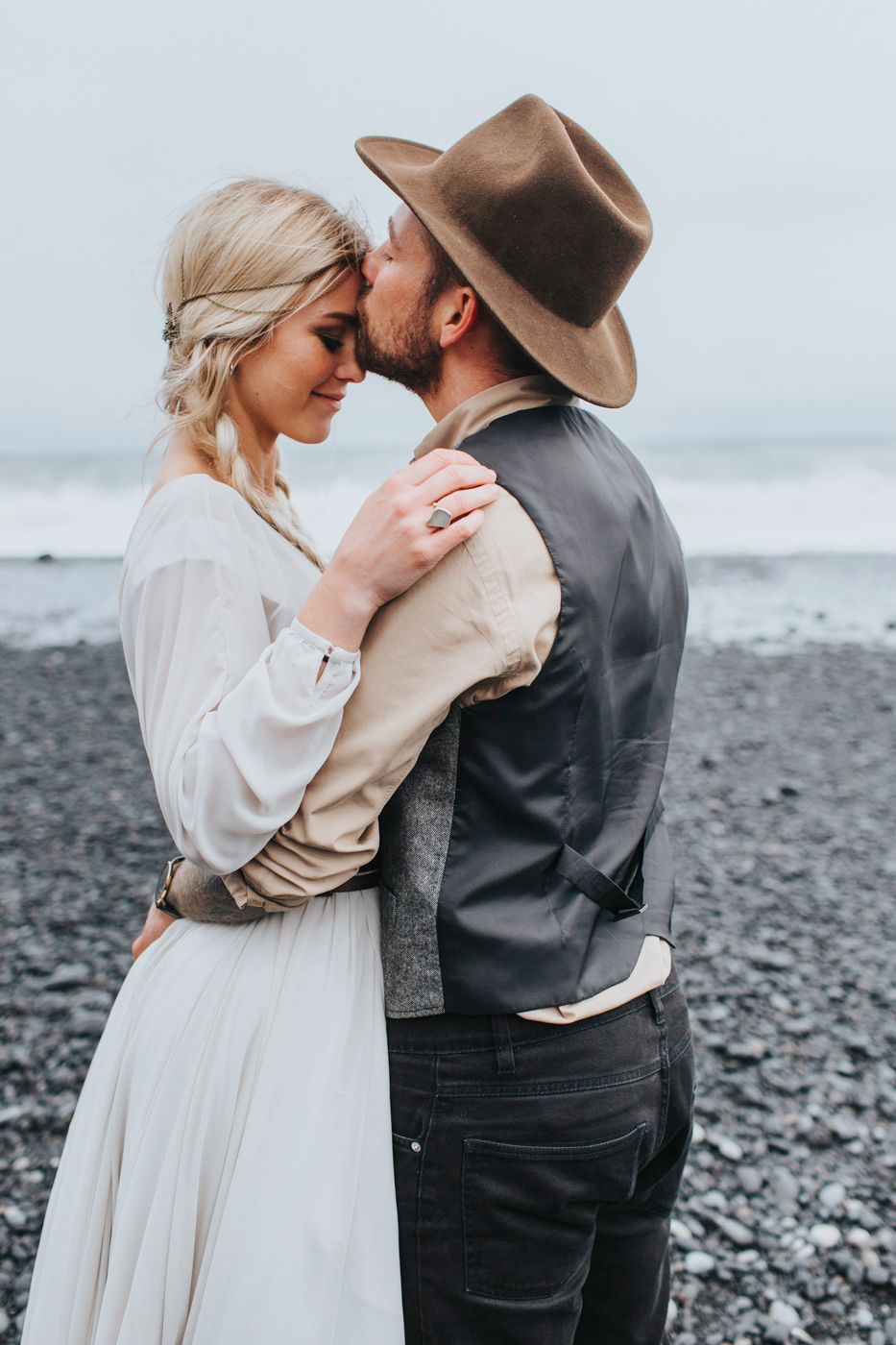 Intimate moment between newlyweds on a misty beach with dark volcanic sand and ocean waves.