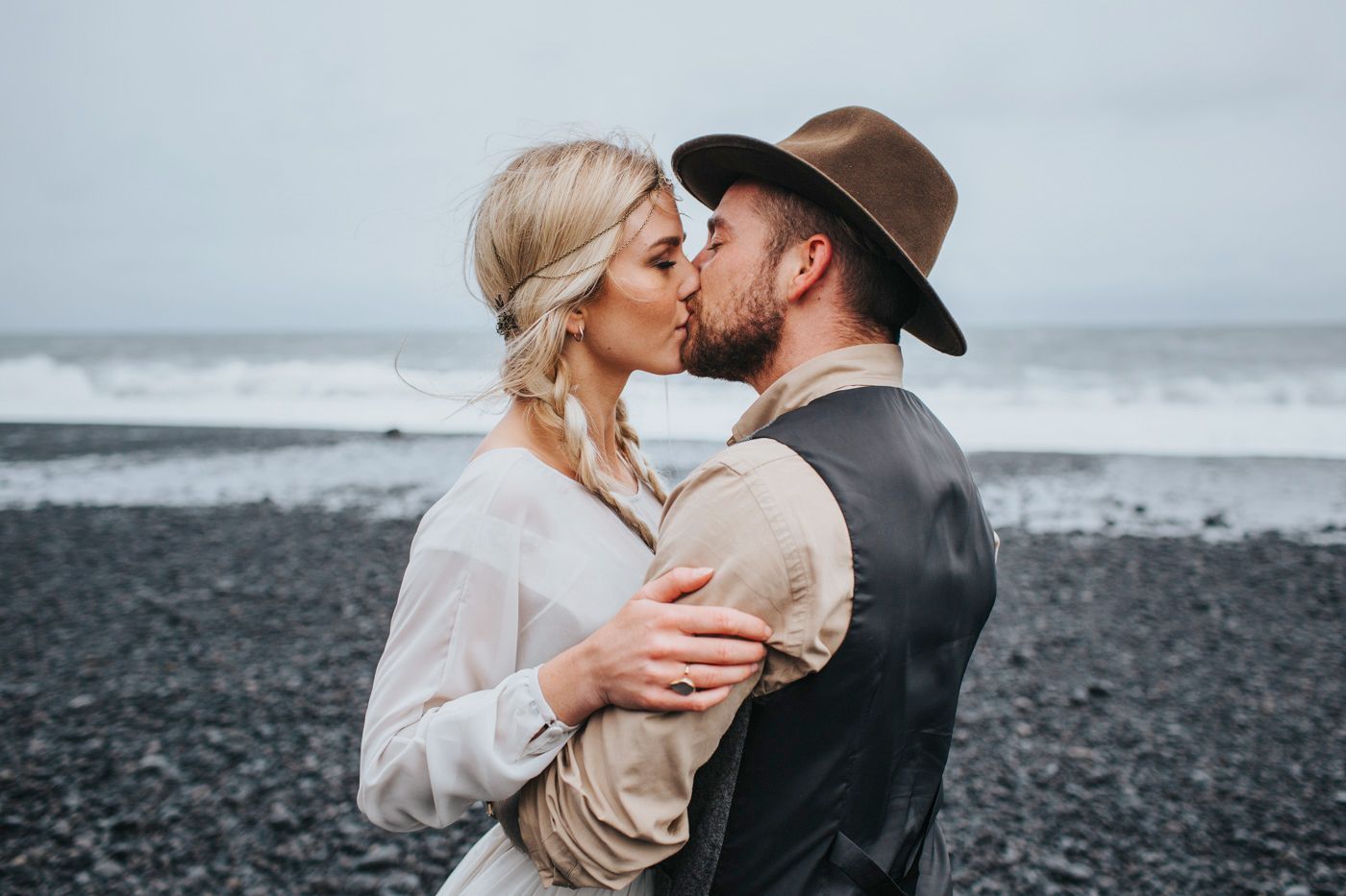 Romantic kiss between newlyweds on moody black sand beach with crashing waves.