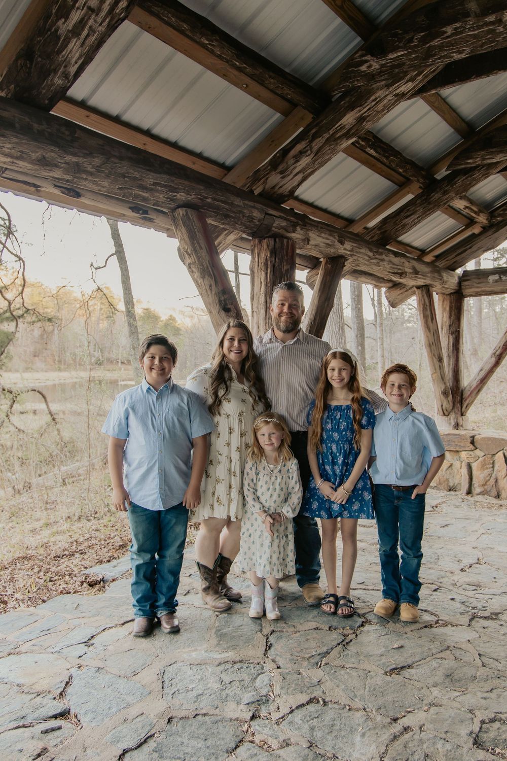 Family of six posing under rustic log shelter in wooded outdoor setting, casual portrait.