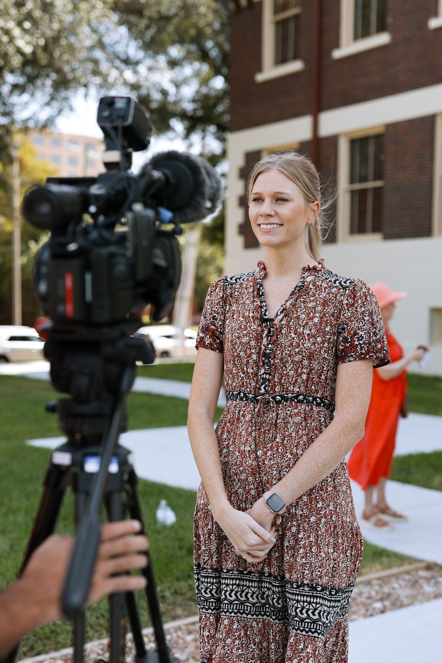 A woman in a floral print dress stands in an outdoor setting being filmed by professional video cameras and crew.