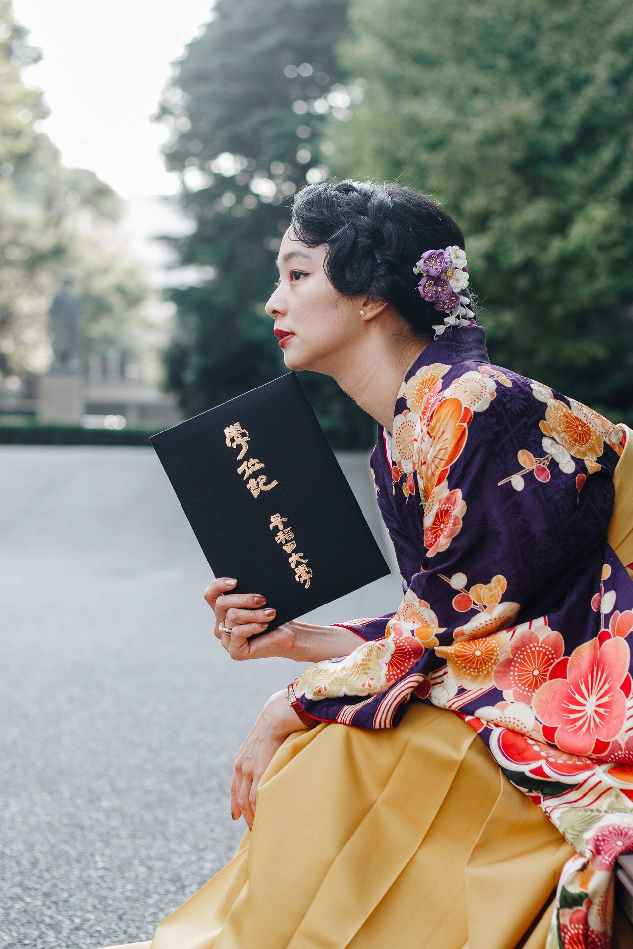 Person wearing a colorful floral kimono holds a black book while posing against a blurred outdoor background.