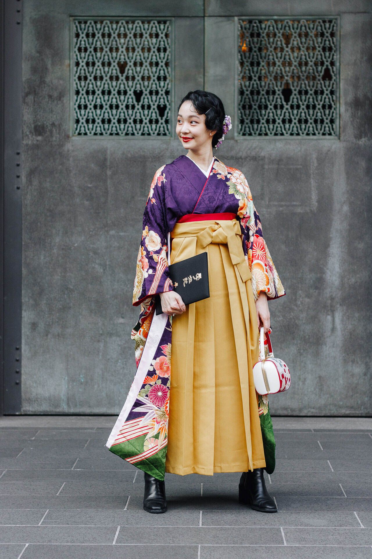 Figure in purple and gold kimono with floral design stands against decorative wall pattern.