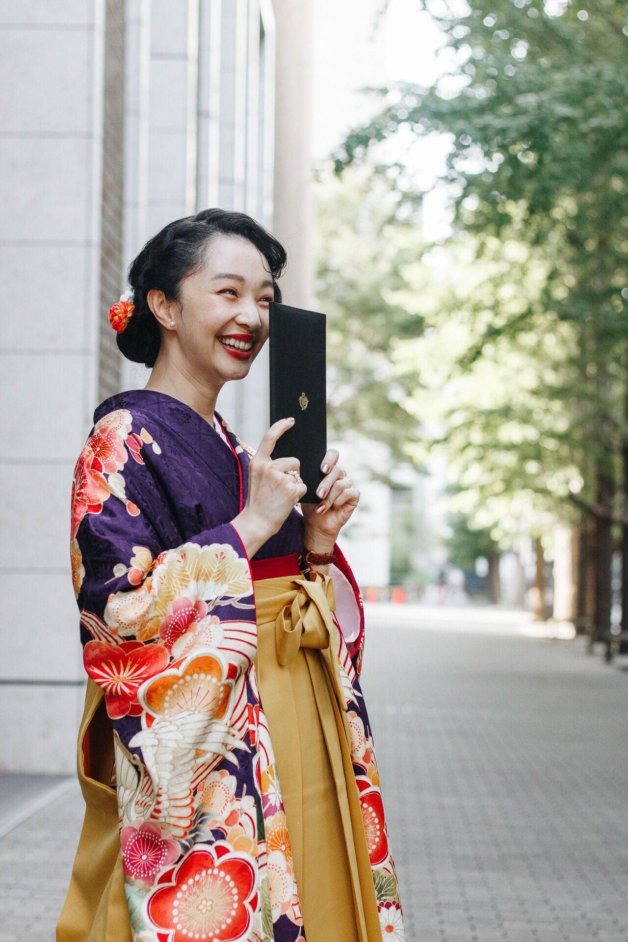 Person in vibrant kimono holds black book while smiling outdoors near building.
