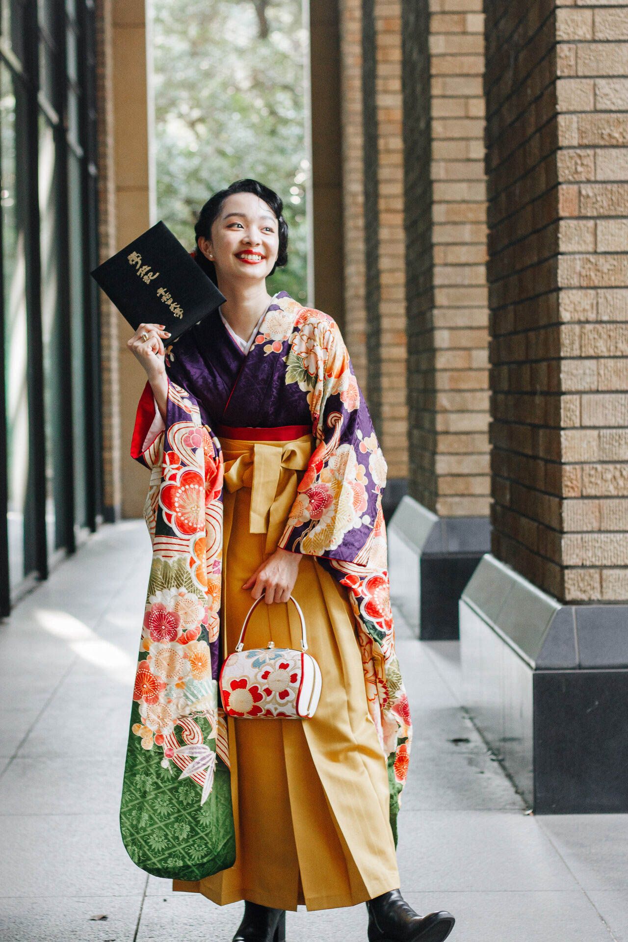 Person wearing floral kimono poses with black book and green bag against brick building.
