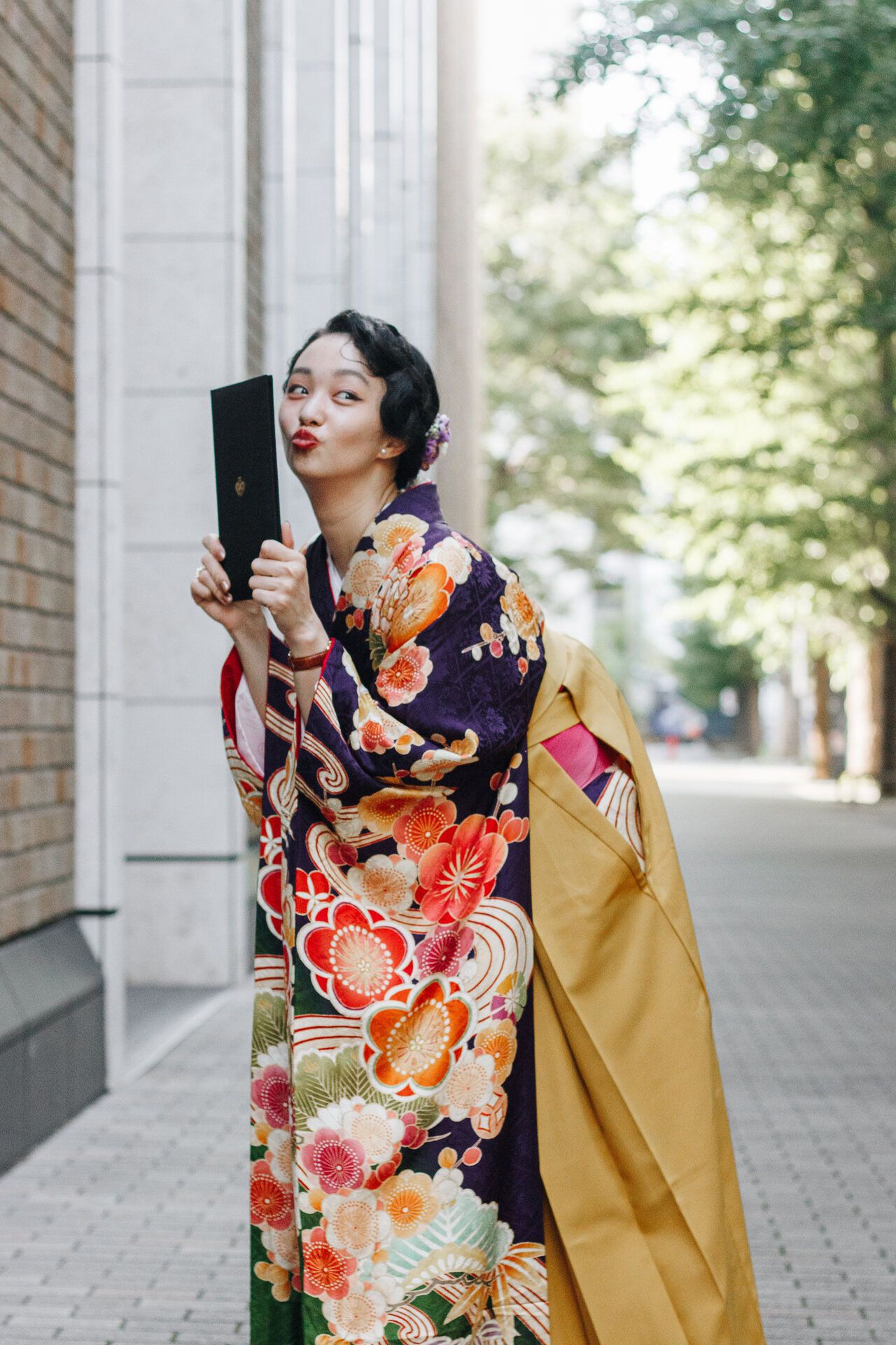 Person in traditional Japanese dress poses with black book against architectural backdrop.