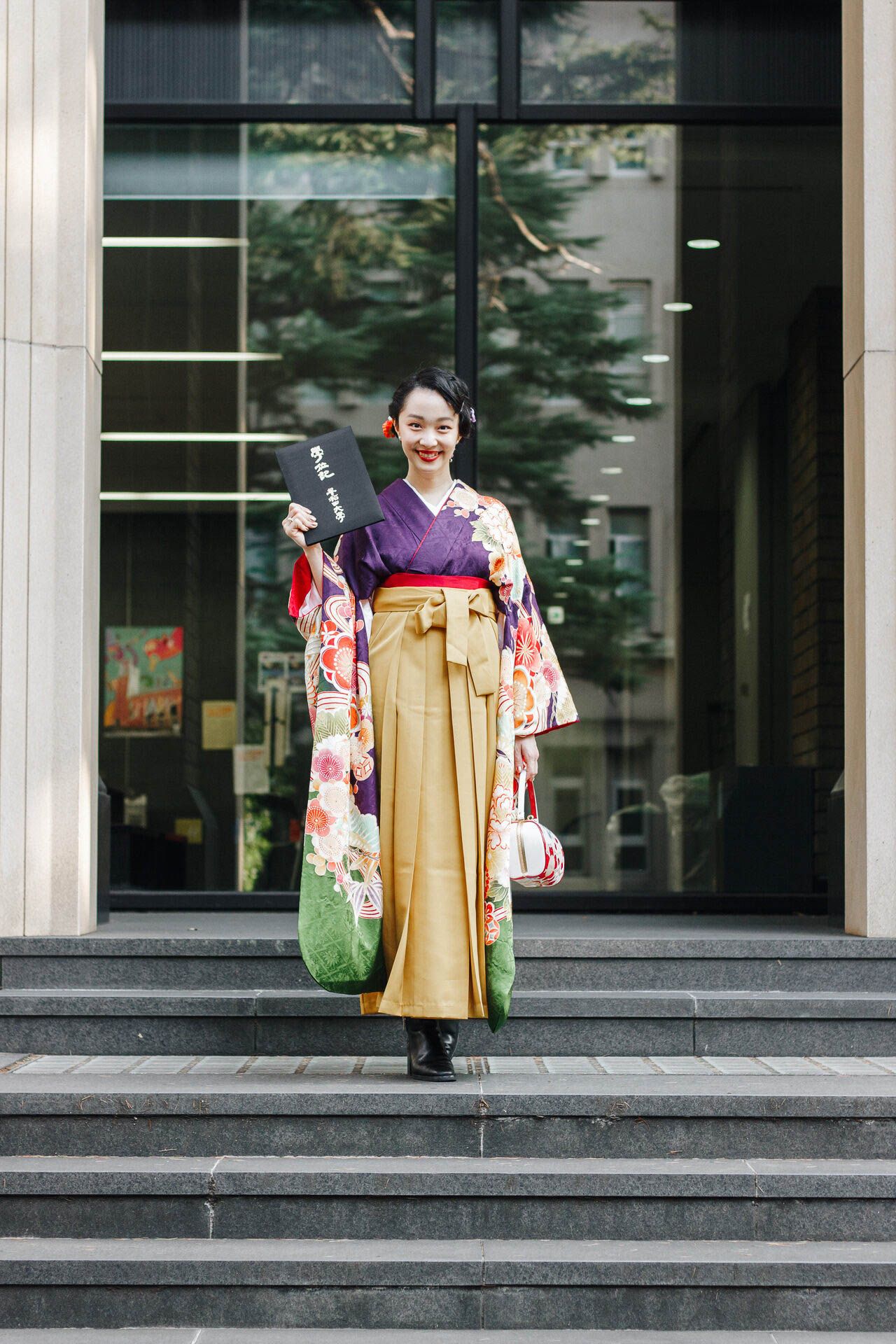 Person in purple and gold kimono stands on steps holding a black book and green bag.