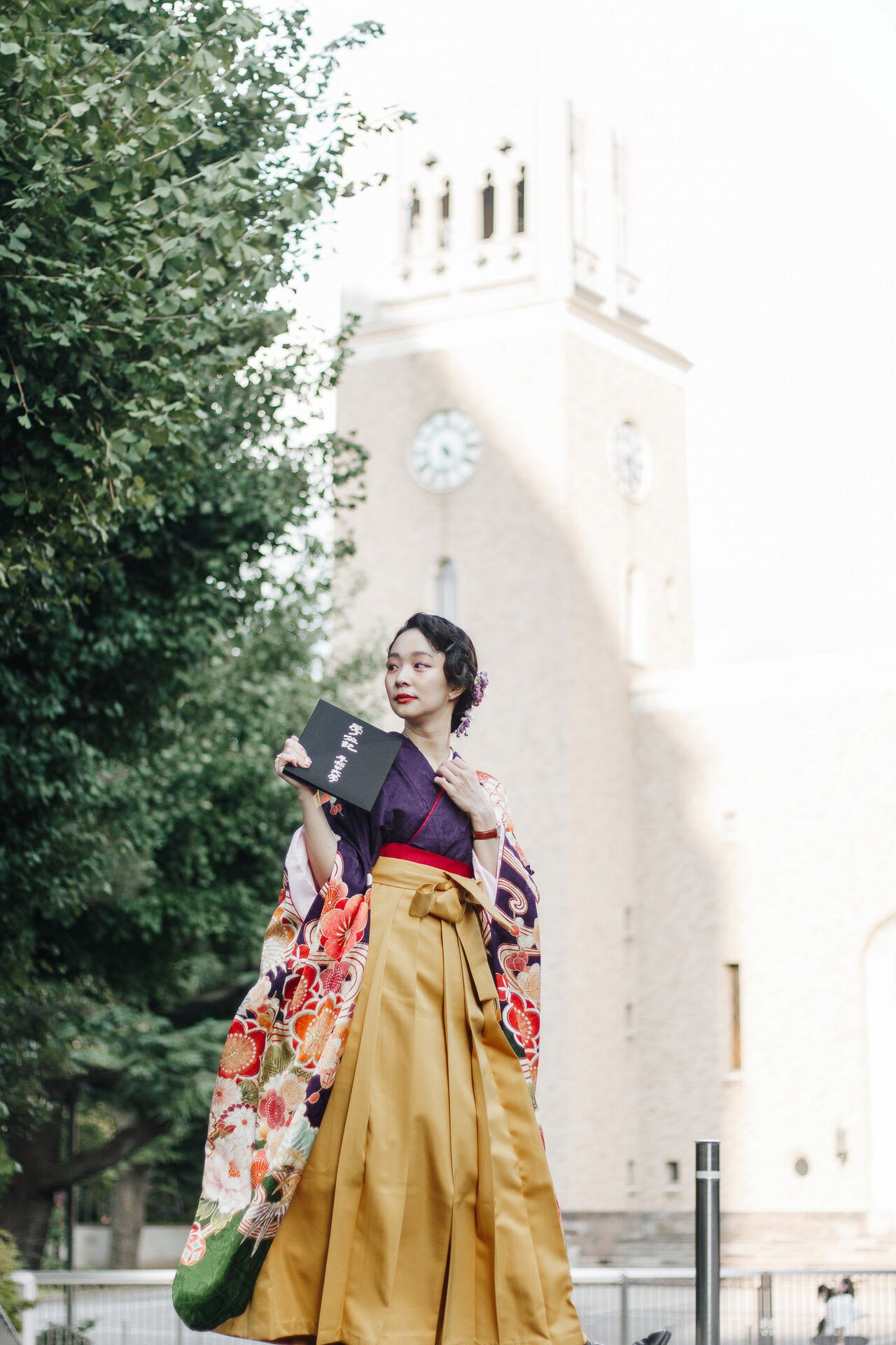 Figure in kimono poses with book in front of historic clock tower building.