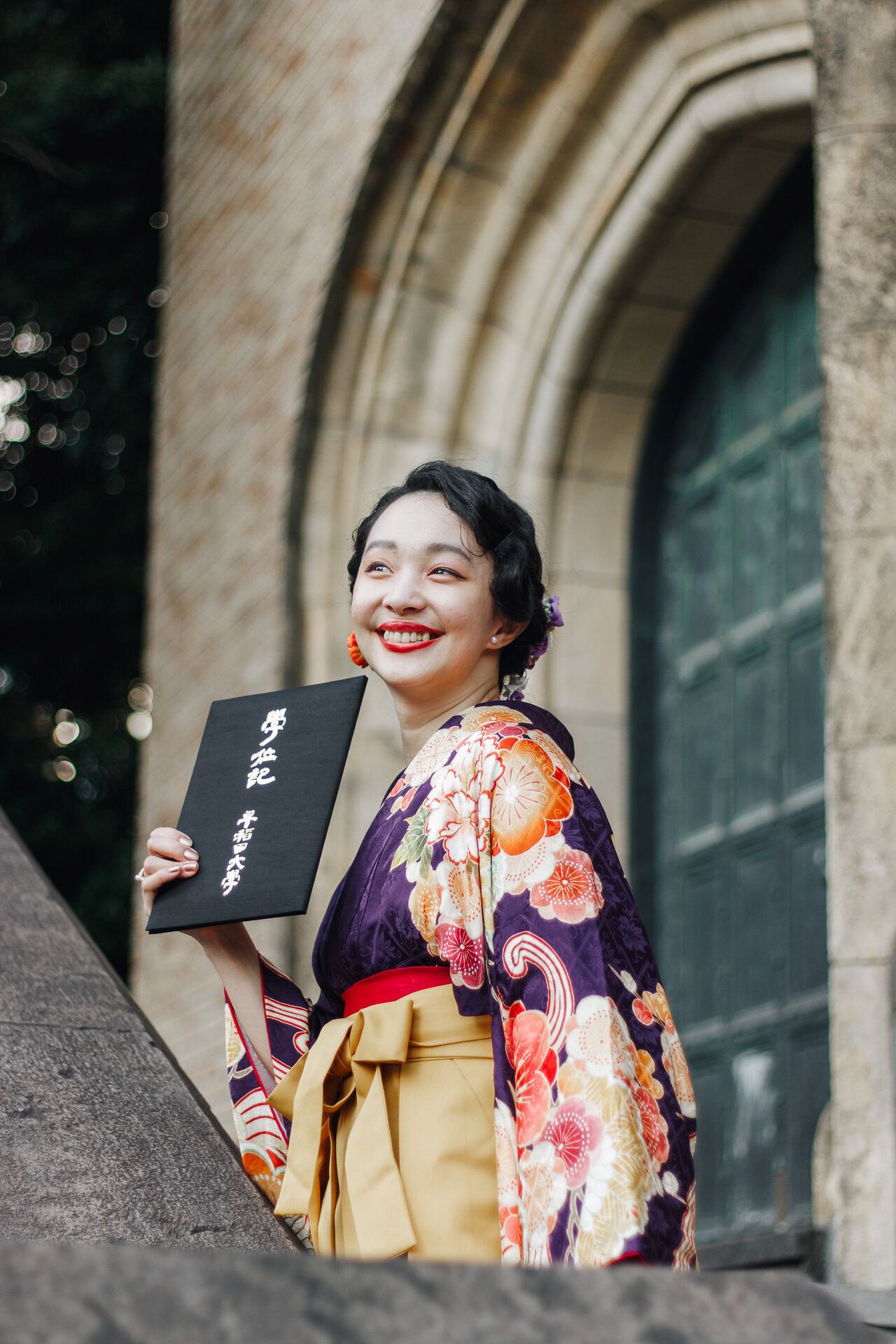 Person in traditional Japanese attire smiles while holding a black book near stone architecture.