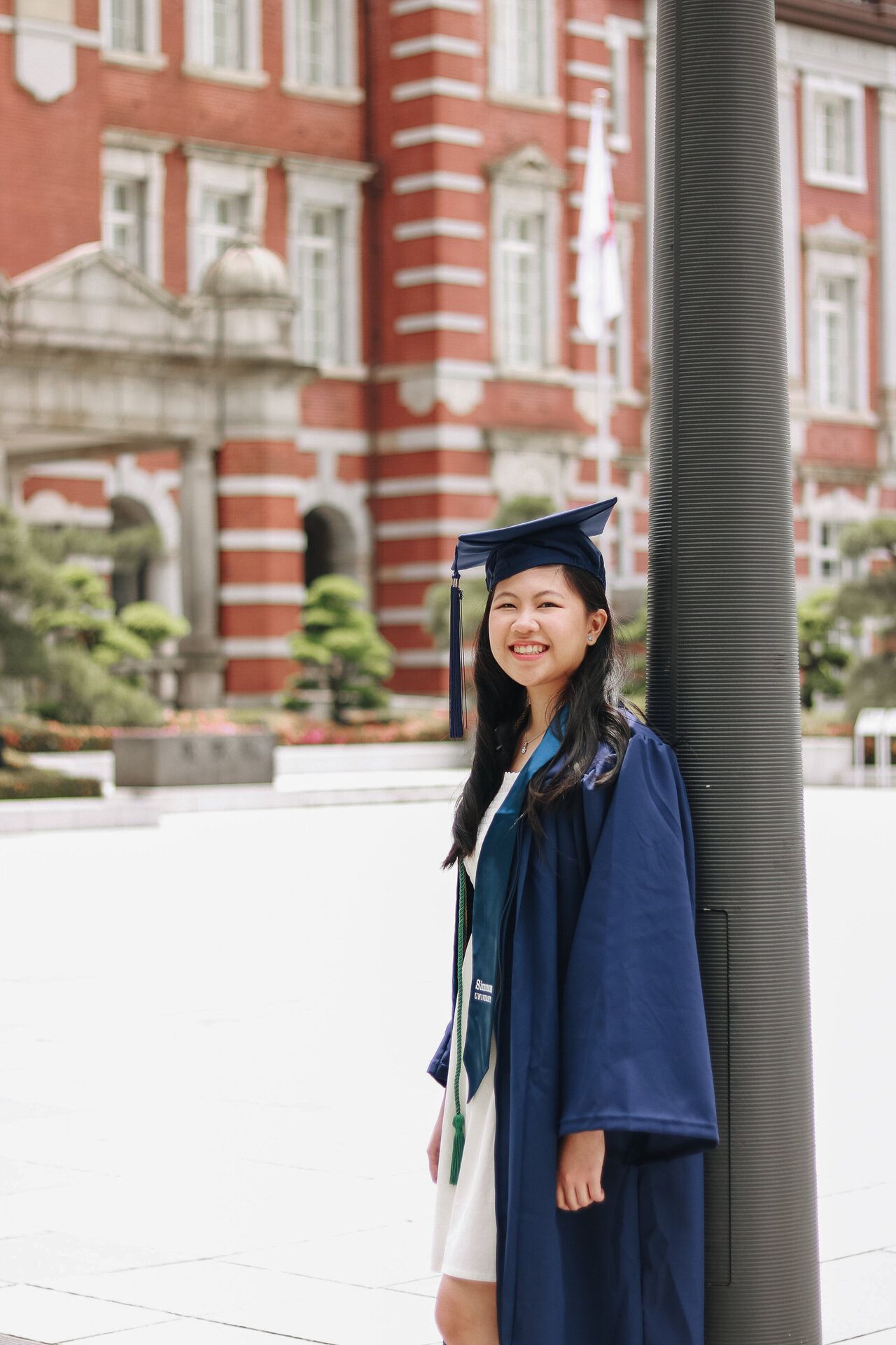 Graduate in navy blue academic regalia poses in front of red brick university building.