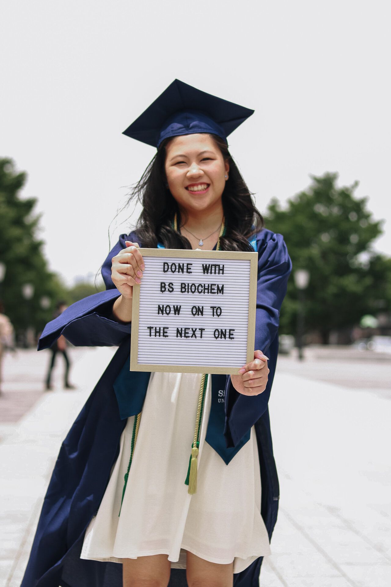 Graduate in blue cap and gown holds letter board with message about completing biochemistry degree.